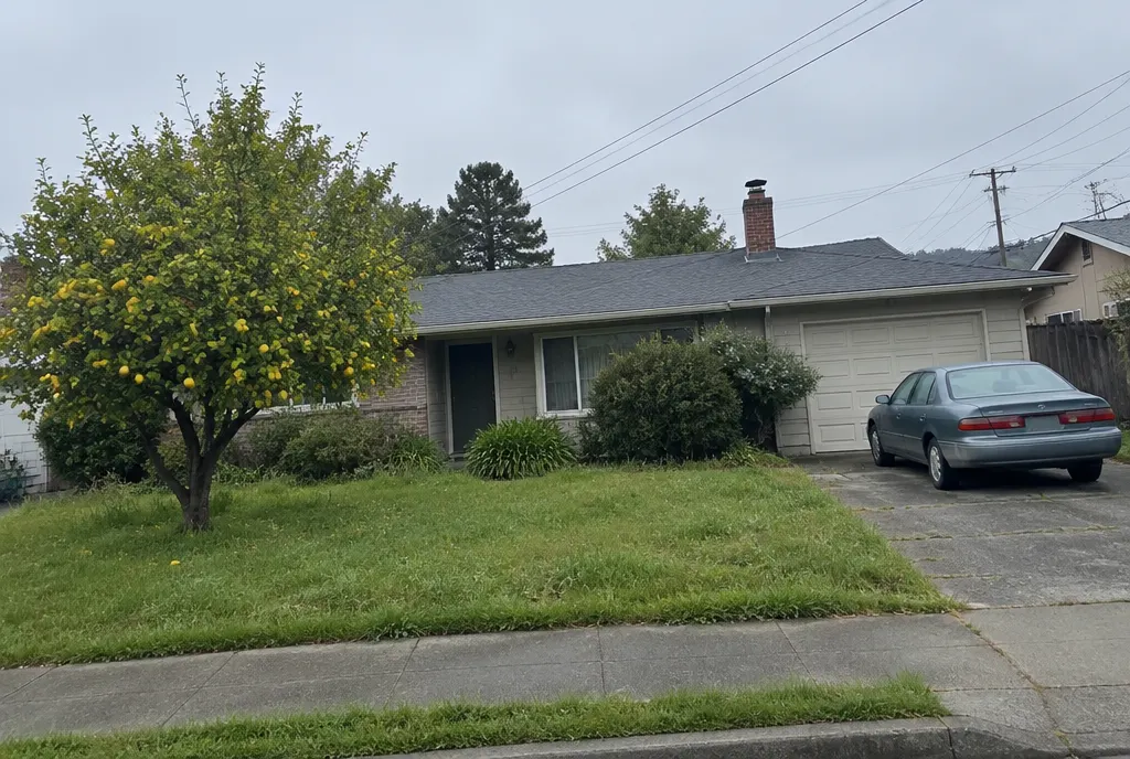 An older single story ranch house with a lemon tree in the front yard and a sedan in the driveway on an overcast morning